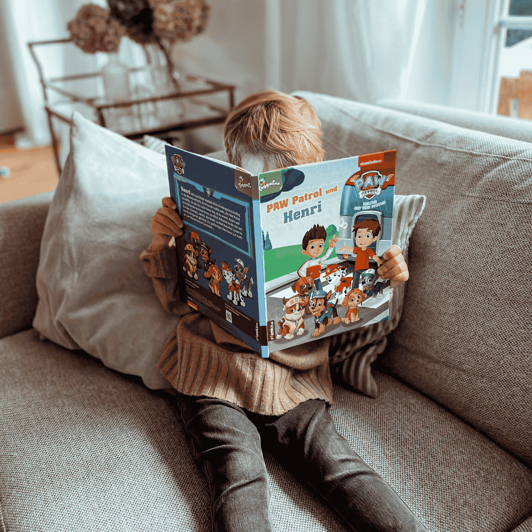 Ein kleines Kind sitzt auf einer Couch und liest ein "PAW Patrol"-Buch, sein Gesicht ist hinter dem Einband verborgen. Der Raum ist mit weichem, natürlichem Licht erfüllt.